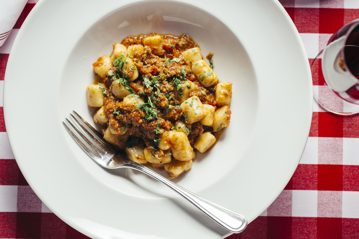 overhead photo of gnocchi bolognese with a fork sitting on the lip of the plate on a red and white checkered table cloth