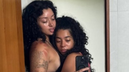 Duas mulheres de cabelos escuros abraçadas tirando uma selfie em um espelho