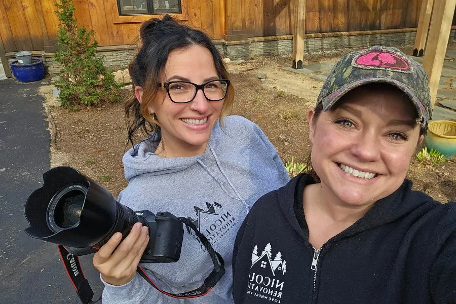 Two women, one wearing glasses and a gray hoodie, the other in a camouflage cap and black hoodie, smiling for a selfie with a professional camera, standing outside with a wooden fence and greenery in the background.


