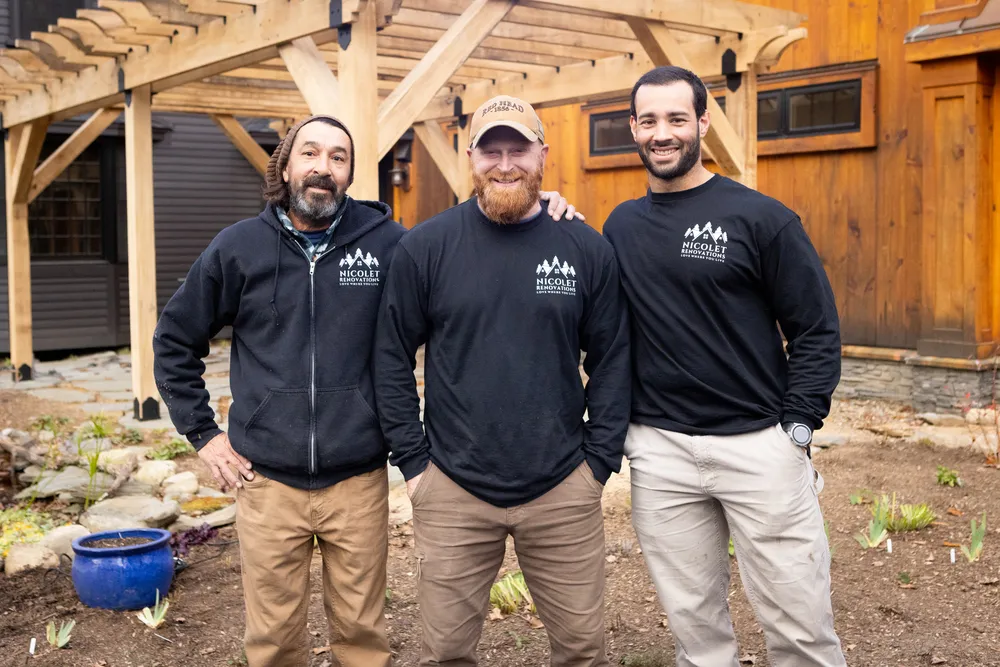 Three men, each wearing a black hoodie with the "Nicolet Renovations" logo, are standing confidently in front of a wooden structure with a stone foundation, smiling and posing for the photo.