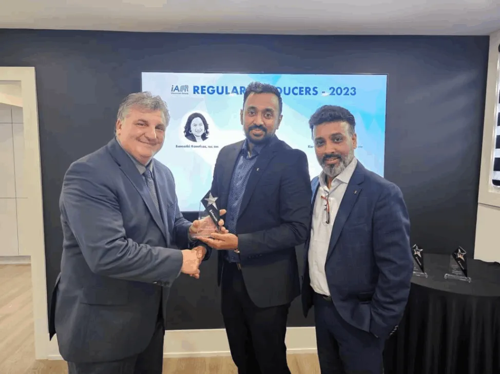 Three men in business suits stand together in a corporate setting, with one man handing an award to another. A digital screen in the background displays the text "Regular Producers - 2023." The award recipient smiles as he receives the trophy, symbolizing recognition for outstanding performance. The setting suggests a professional awards ceremony or corporate event. Toronto, Canada