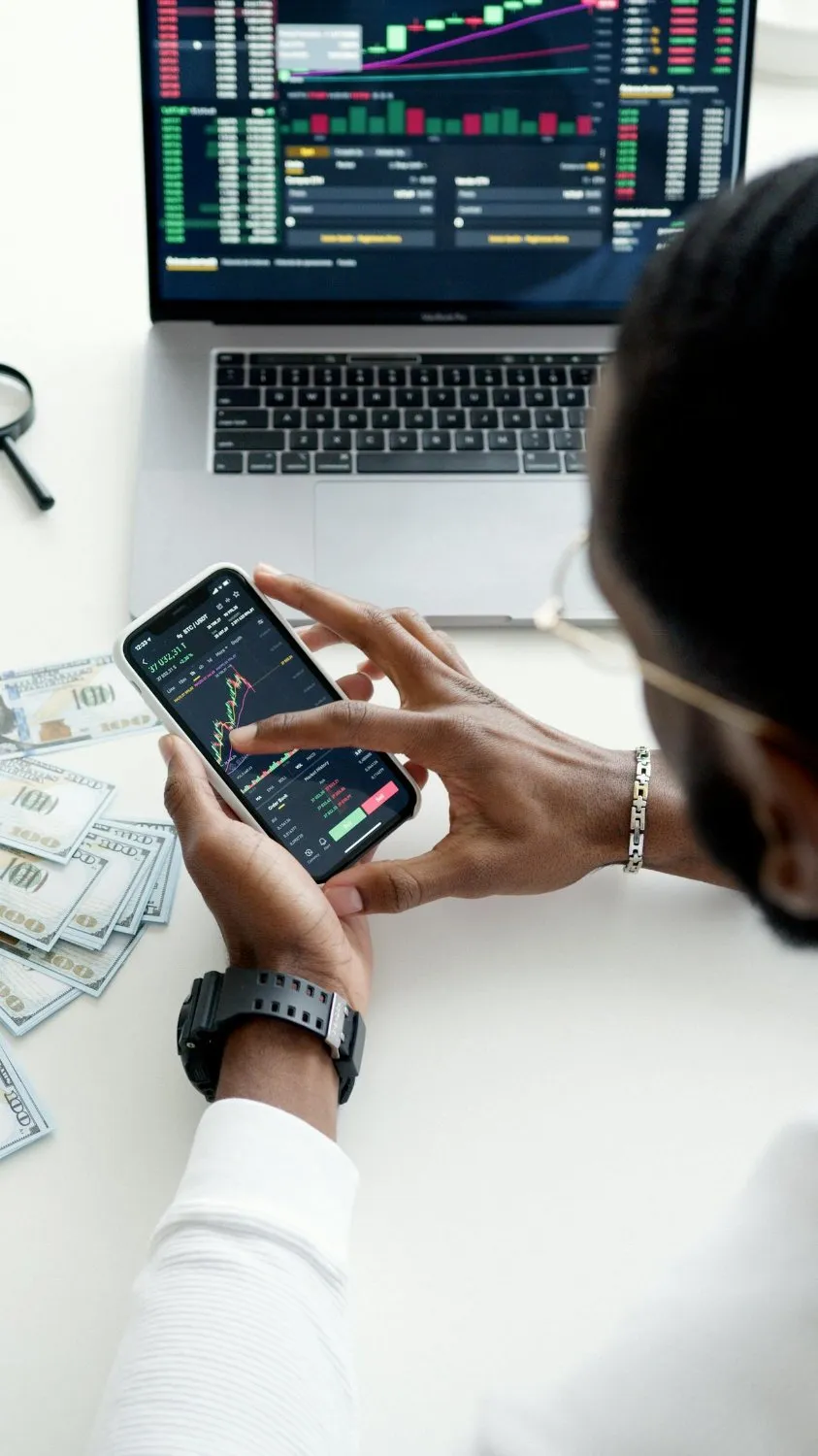 Business person reviewing investment performance on a smartphone, with financial growth charts on a laptop in the background. Toronto, CA