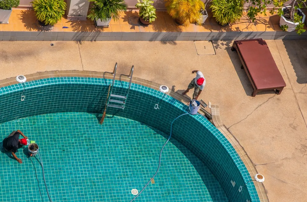 Two workers clean the floor of an empty tiled swimming pool using hoses and buckets.