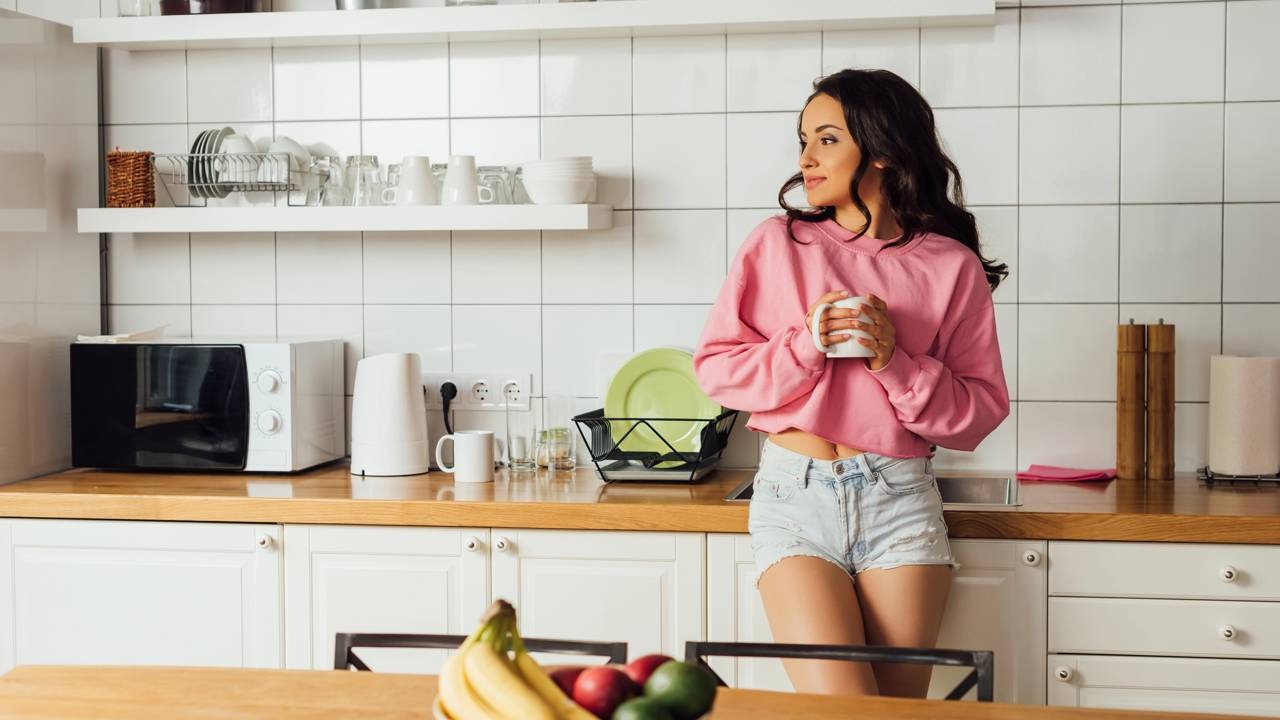Prepare seu shot matinal na cozinha, uma dose diária para impulsionar a saúde e o bem-estar do sistema imunológico. Créditos: depositphotos.com / AndrewLozovyi Mulher sorridente com caneca em cozinha moderna, com micro-ondas e balcão de madeira