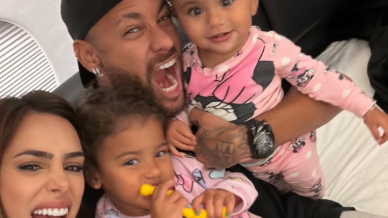 Neymar e Bruna Biancardi em momento descontraído com as filhas Mavie e Mel. Neymar, Bruna Biancardi e duas crianças com pijamas rosa sorrindo em selfie