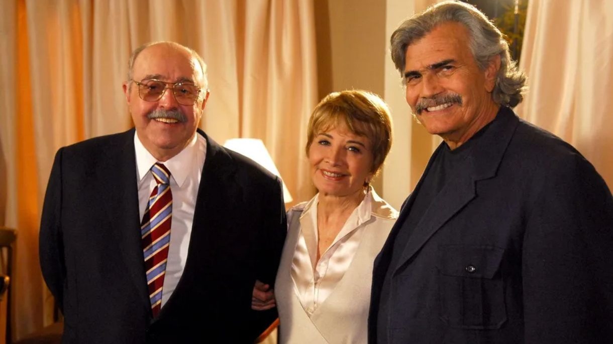 Mulher de cabelo curto loiro entre dois homens, ambos com bigode e sorrindo