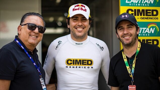 Os irmãos pilotos Cacá e Popó Bueno, filhos de Galvão Bueno, em um dos eventos de Stock Car. Três homens sorrindo em um paddock. Um de óculos, outro com macacão Cimed, e o terceiro de boné