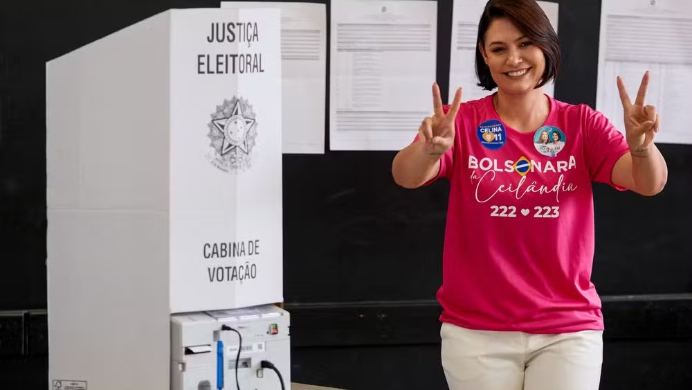 Michelle Bolsonaro sorri, com sinais de vit&oacute;ria, ao lado de cabine e urna eletr&ocirc;nica de vota&ccedil;&atilde;o