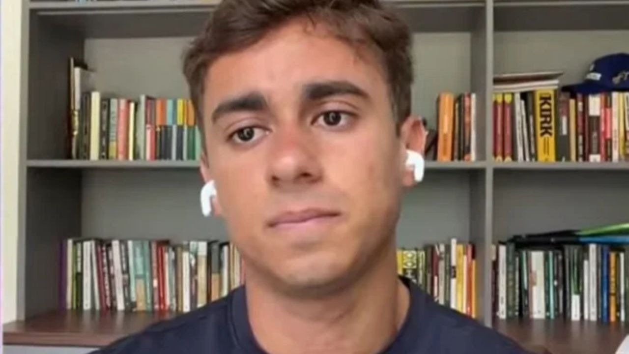 Nikolas Ferreira, eleito deputado federal pelo PL. Jovem com fones brancos, camisa escura, estante de livros ao fundo, livro 'KIRK'