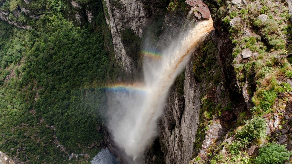 Cachoeira poderosa caindo em cânion rochoso e verde, com arco-íris na névoa.
