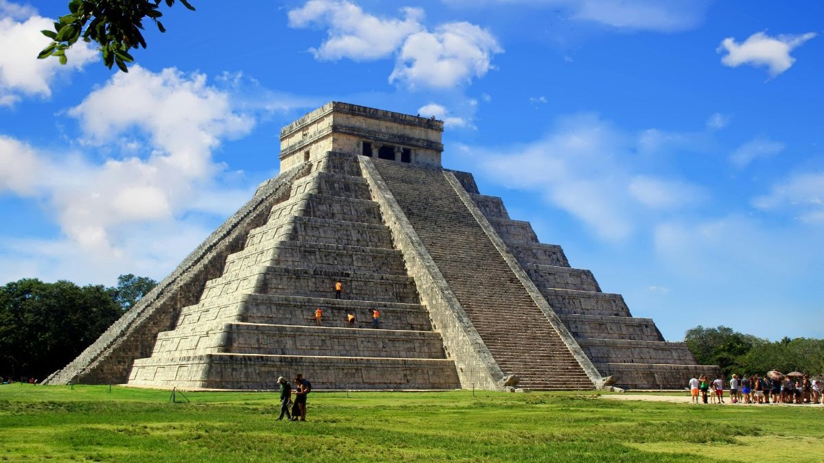 A pirâmide de Kukulcán, símbolo da civilização maia, figura entre as 7 maravilhas do mundo moderno. Pirâmide de Kukulcán em Chichén Itzá sob céu azul com pessoas e grama verde.