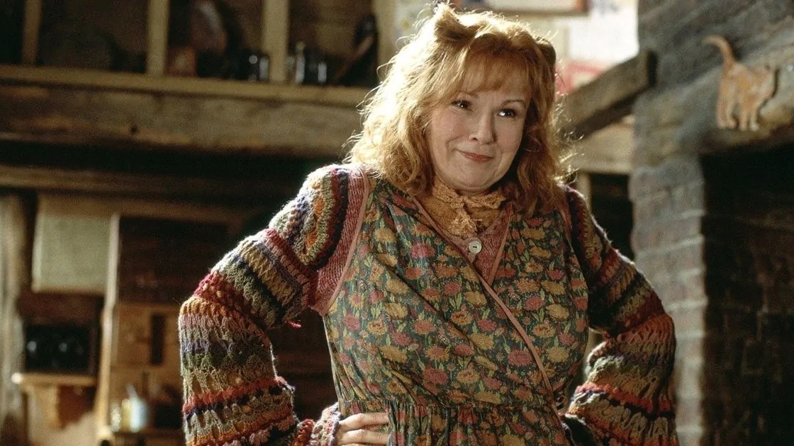 Molly Weasley sorri, com cabelo ruivo e su&eacute;ter colorido, em cen&aacute;rio r&uacute;stico.