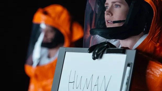 Amy Adams em cena de "A Chegada", um dos filmes de ficção científica sugeridos para uma sessão aconchegante. Amy Adams em traje hazmat laranja segura quadro branco com a palavra "HUMAN".