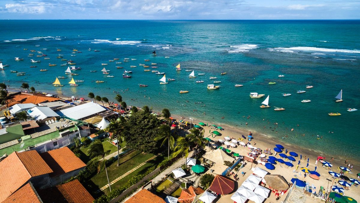 Vista aérea de praia movimentada com mar azul, jangadas, guarda-sóis e edifícios em Porto de Galinhas.