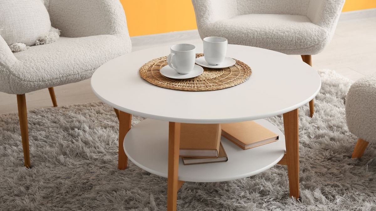 Mesa de centro branca redonda com copos, rodeada por poltronas claras e tapete felpudo cinza.
