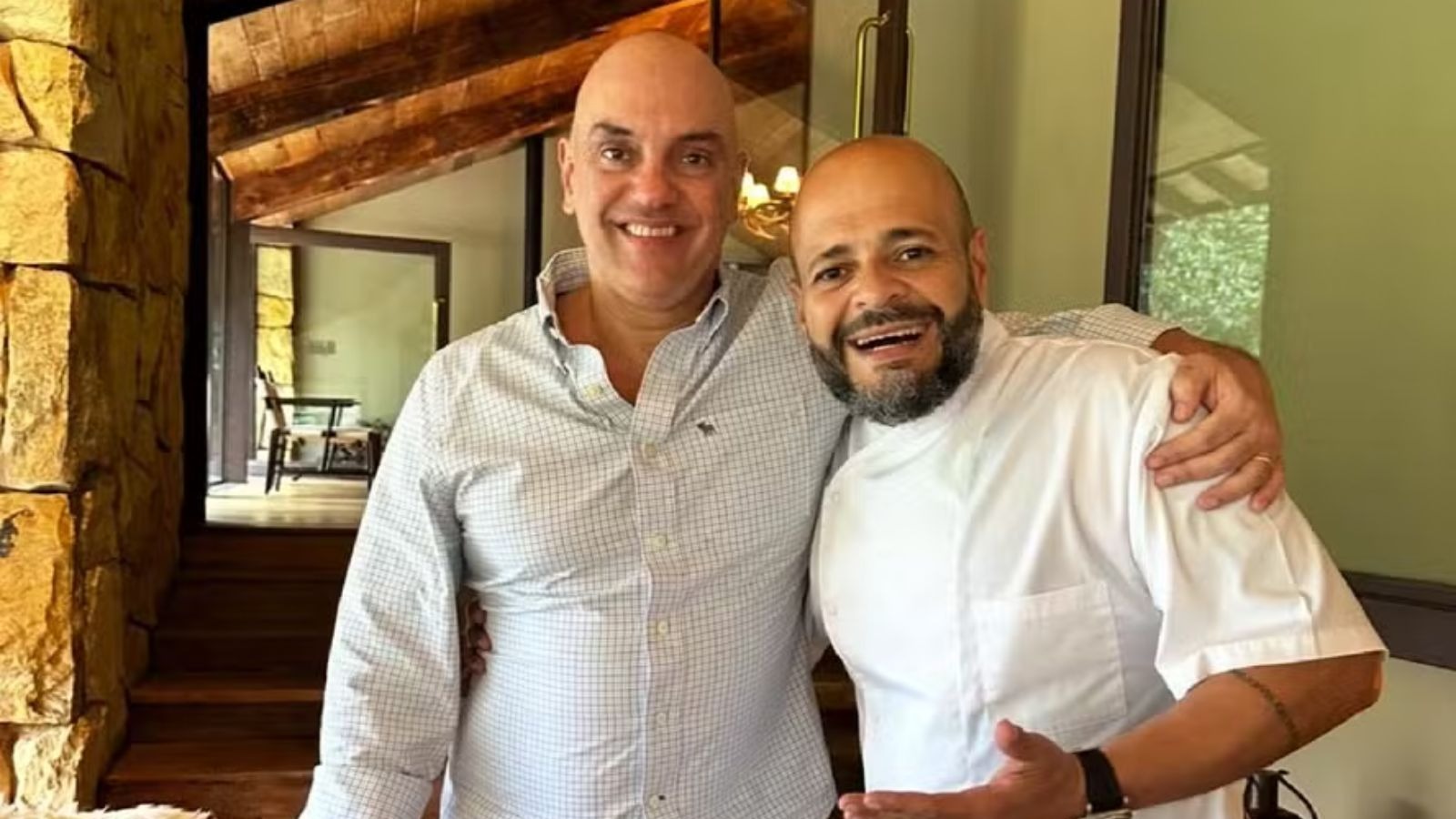 Dois homens carecas sorrindo e abra&ccedil;ados, um de camisa xadrez, outro com barba e jaleco branco de chef