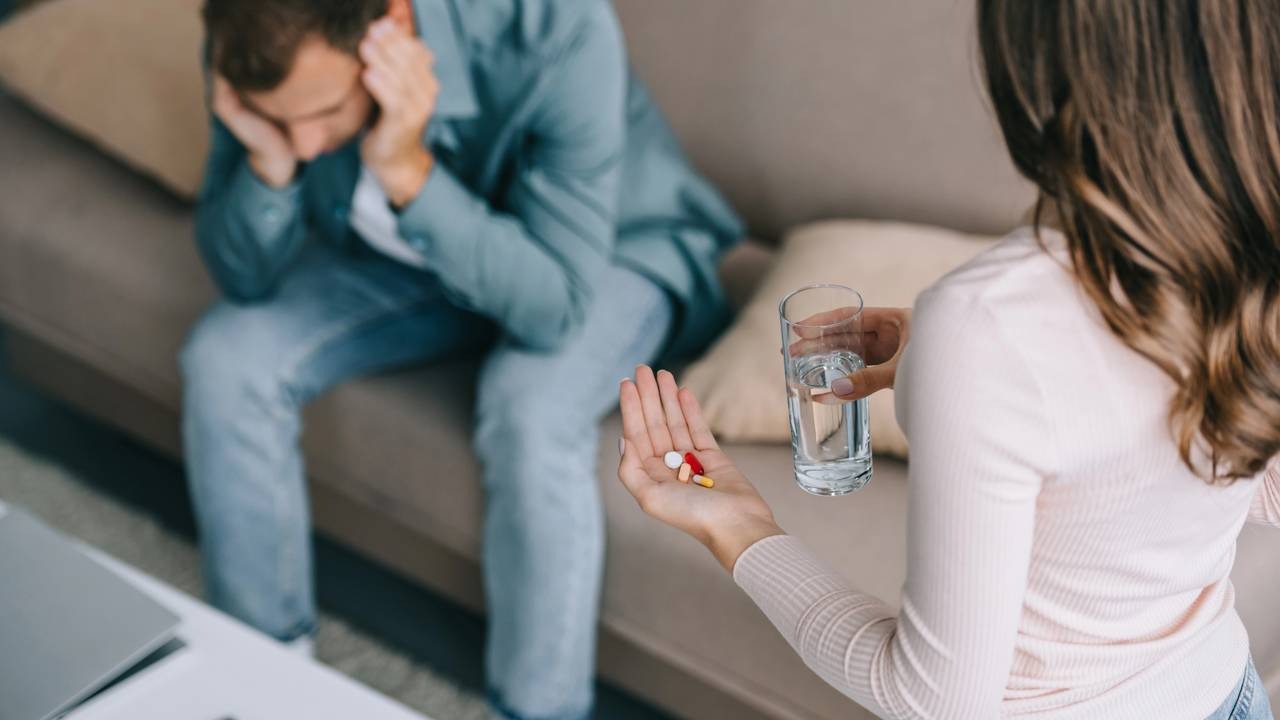 O mal-estar causado pelas mudanças climáticas pode levar à necessidade de cuidado e alívio para os sintomas. Mulher oferece copo de água e comprimidos a homem cabisbaixo no sofá.
