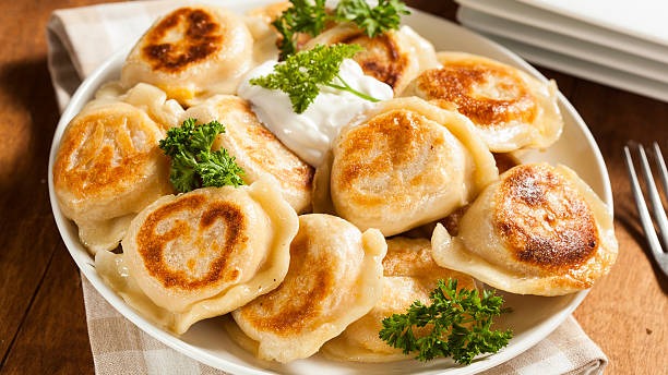 Pierogi, prato tradicional no Paraná, reflete a forte influência da imigração europeia na rica culinária local. Prato branco cheio de pierogi dourados, com salsa e uma porção de creme branco no centro.
