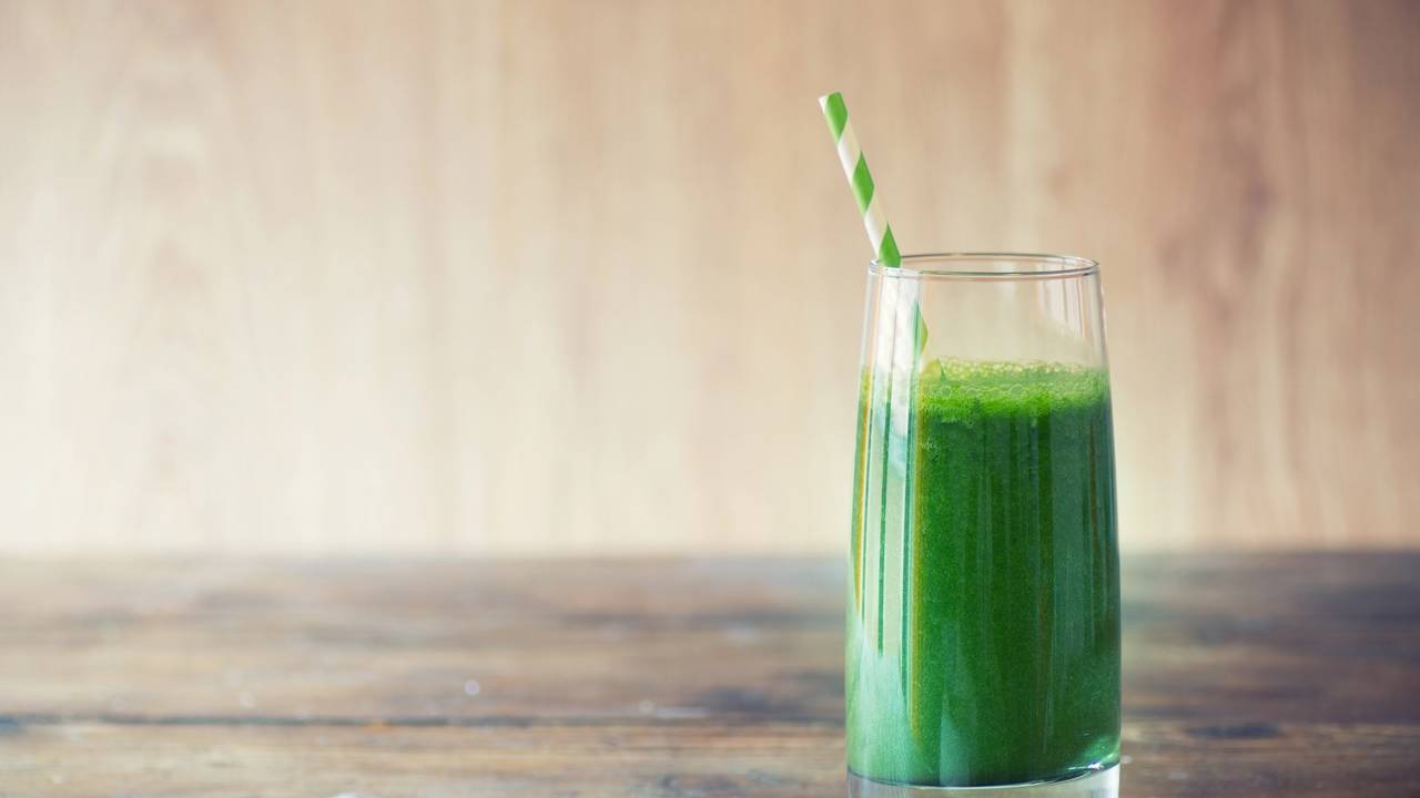Desfrute do suco verde funcional, uma dose matinal de energia e nutrientes essenciais para sua rotina diária. Copo alto de suco verde com canudo listrado em mesa de madeira, fundo claro.