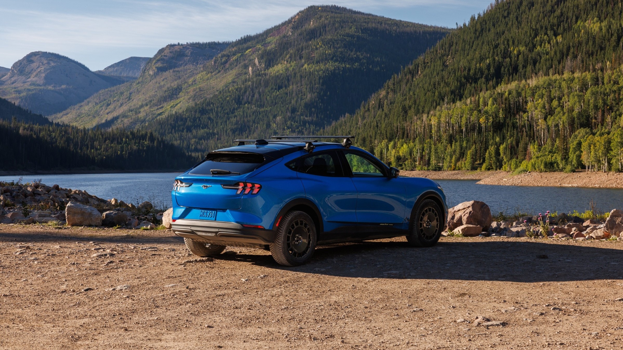 Ford Mustang Mach-E azul, com rack de teto, em estrada de terra pr&oacute;ximo a um lago e montanhas