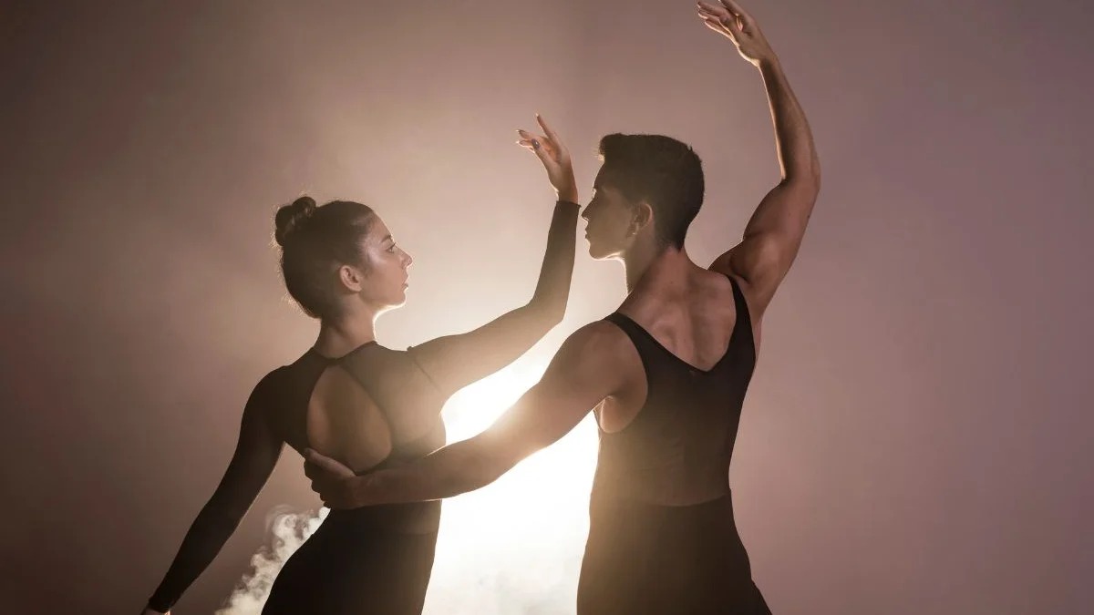 A dança, retratada em pares, é uma atividade completa que une expressão artística e exercício físico com grandes benefícios para a saúde Duas pessoas dançando, de perfil, vestindo roupas pretas, com luz forte e fumaça