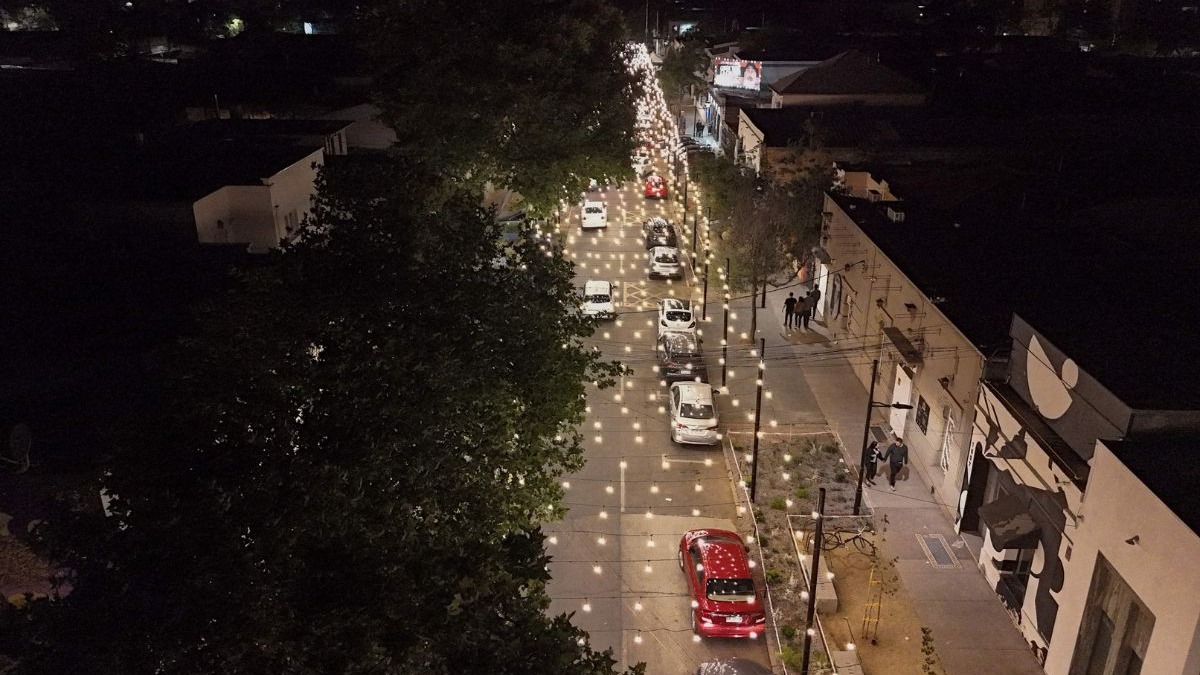 Vista aérea noturna de uma rua arborizada com carros e pessoas, iluminada por luzes penduradas.