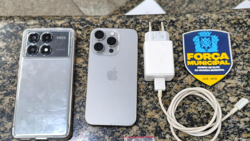 Celulares apreendidos pela Força Municipal durante a prisão dos suspeitos de furto de motocicleta no Rio - Foto; Reprodução Dois smartphones (Poco e iPhone), carregador, cabo e brasão da Força Municipal em mesa