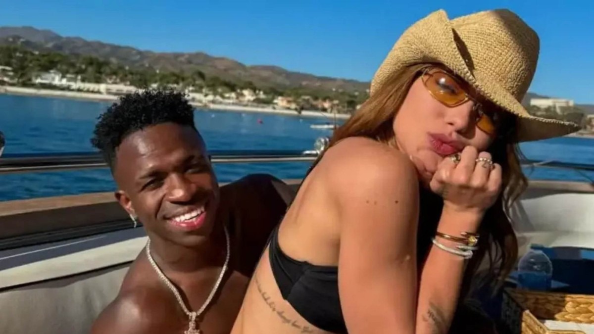Vini Jr. sorrindo e mulher de chap&eacute;u e &oacute;culos em um barco com mar ao fundo