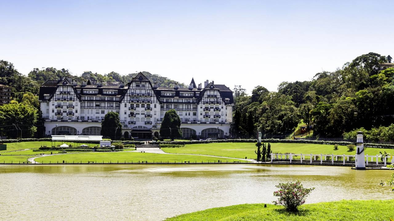 Destinos de serra no Brasil encantam com arquitetura europeia e paisagens idílicas, ideais para as férias de julho. Grande hotel estilo europeu às margens de um lago, com gramado e montanhas arborizadas.
