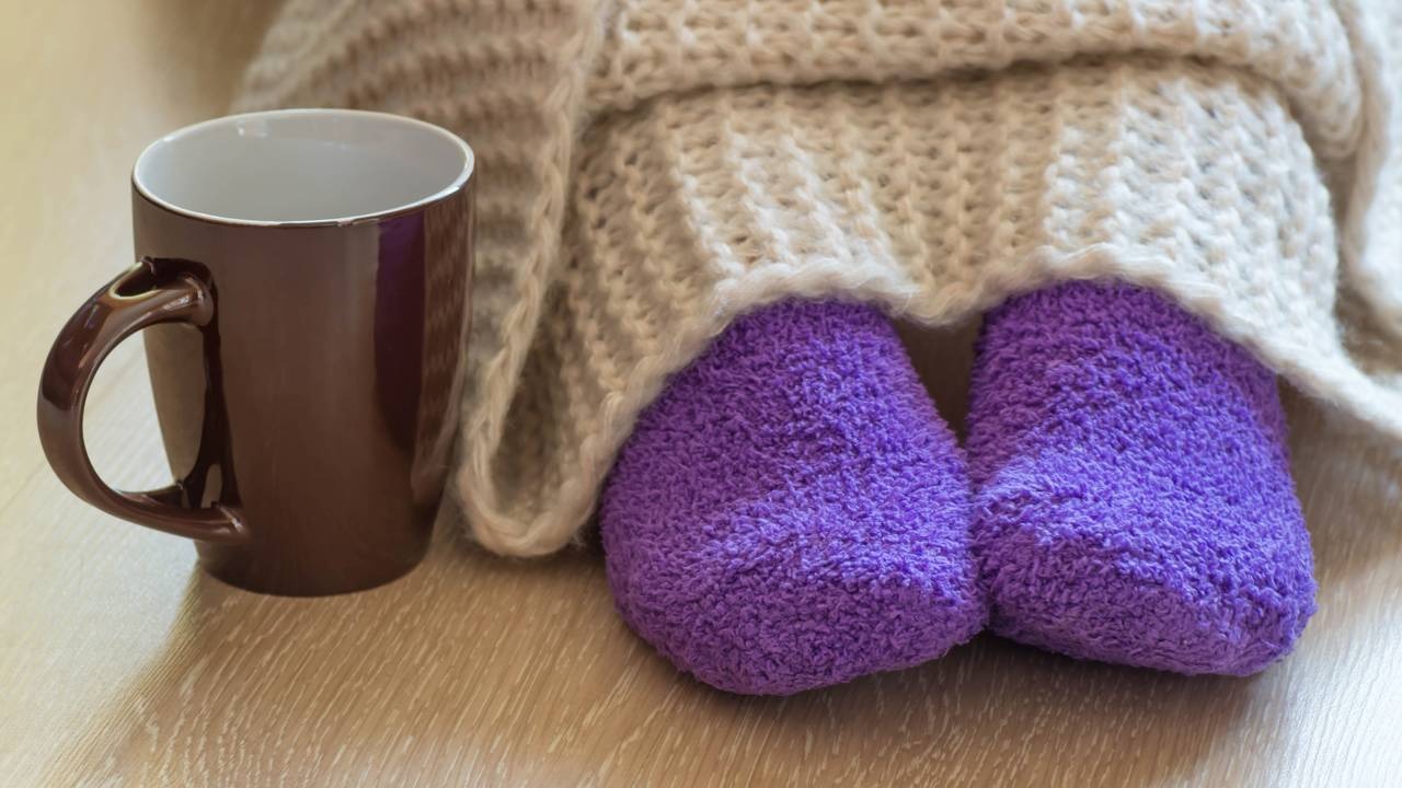 Combata o frio com hidratação e aconchego para proteger a garganta e a imunidade. Caneca marrom, pés com meias roxas macias e cobertor claro sobre piso de madeira.
