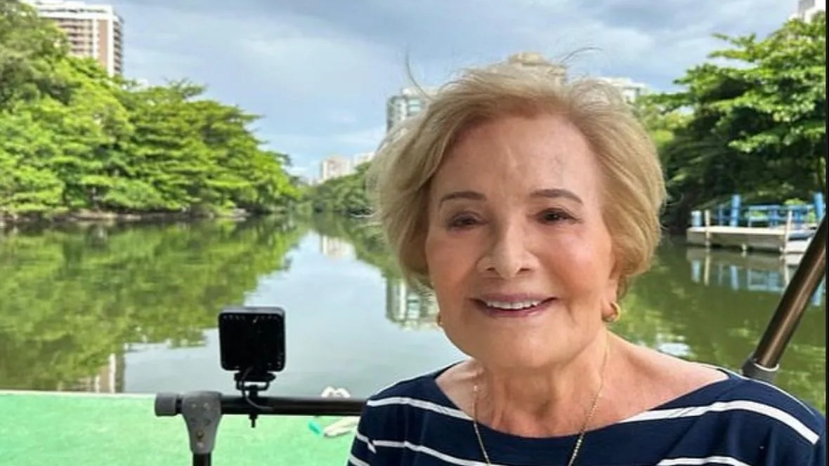 A consagrada atriz Glória Menezes em um registro que destaca sua simpatia e sorriso marcante. Foto: Reprodução Glória Menezes sorrindo, cabelo loiro, blusa listrada azul e branca, rio e árvores ao fundo
