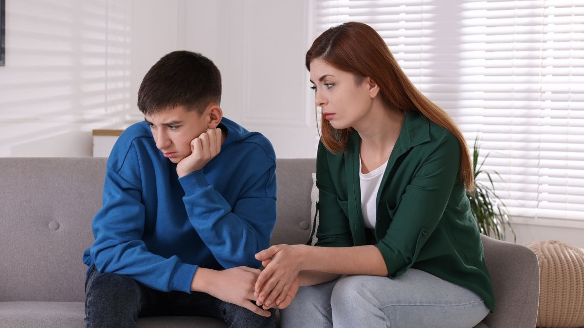Jovem triste sentado no sofá, com uma mulher adulta olhando para ele com preocupação.