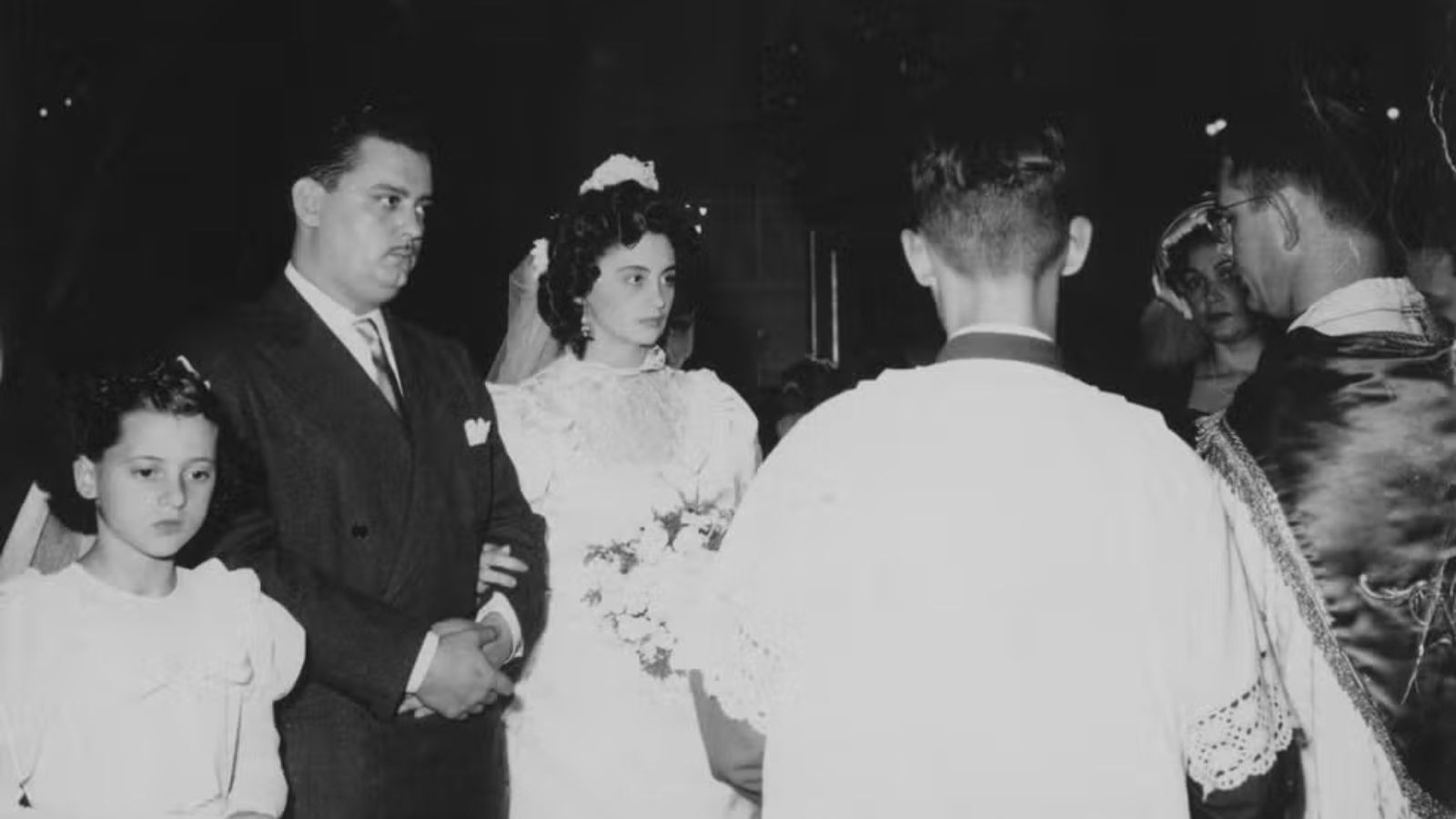 Noiva e noivo de terno em casamento, menina e padre de costas, foto preto e branco