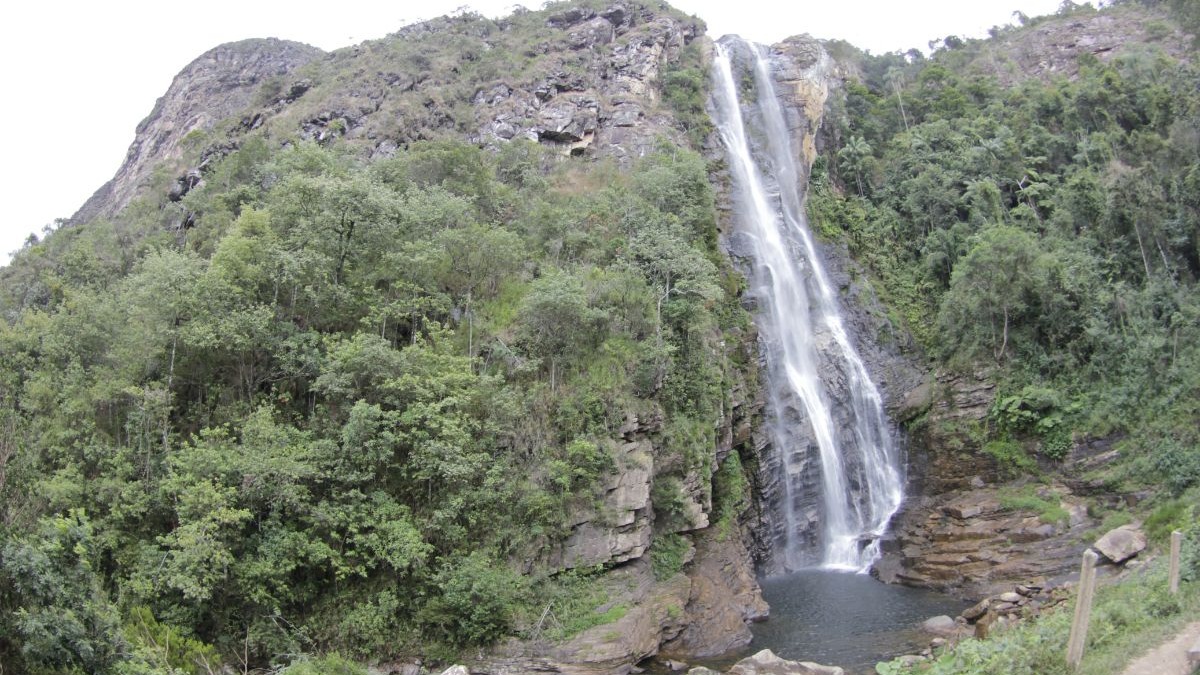 Cachoeira alta e estreita caindo de uma montanha rochosa coberta por vegeta&ccedil;&atilde;o verde exuberante.