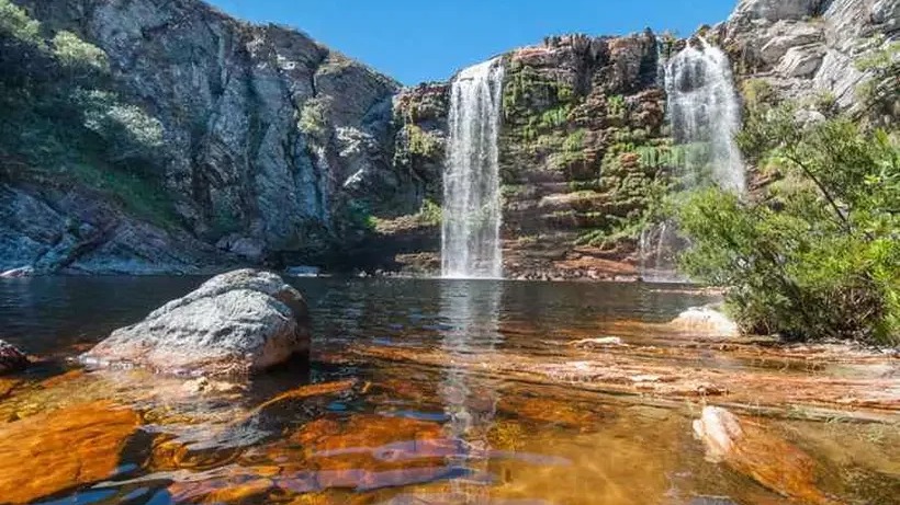 A exuberância da Serra do Cipó, em Minas Gerais, é um convite para explorar cachoeiras e paisagens naturais. Cachoeira alta cai em lago límpido, rodeado por paredões rochosos e vegetação verde.