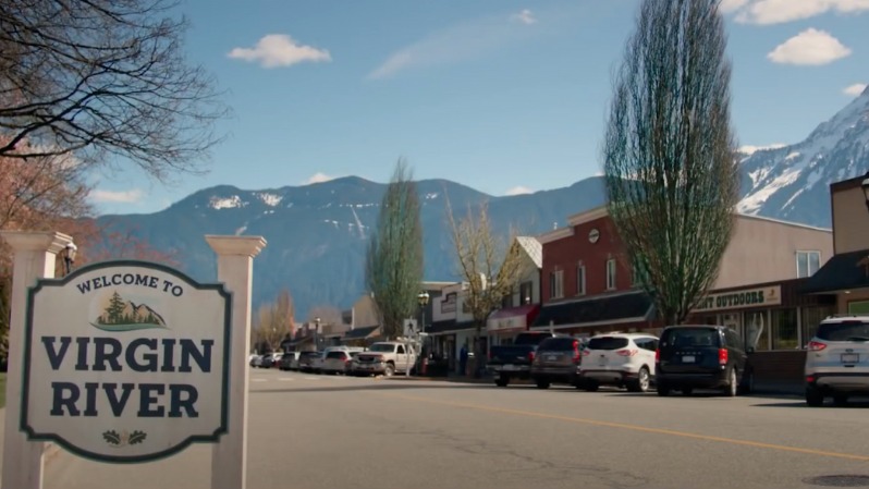 O icônico letreiro de "Virgin River" em uma das locações canadenses que compõem a cidade fictícia da série. Placa "Welcome to Virgin River" na beira de uma rua com carros e montanhas nevadas.