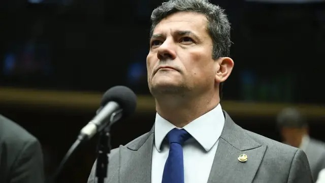 Sergio Moro, um dos governadores eleitos pelo PL nas eleições de 2026. Homem de terno cinza, camisa branca, gravata azul, com microfone e pin na lapela, olhando para cima