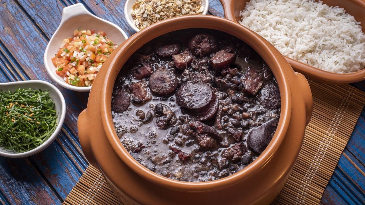 Feijoada completa com os acompanhamentos tradicionais, como arroz, couve e farofa, um prato clássico da culinária brasileira. Panela de barro com feijoada, arroz, farofa, vinagrete e couve refogada em mesa rústica