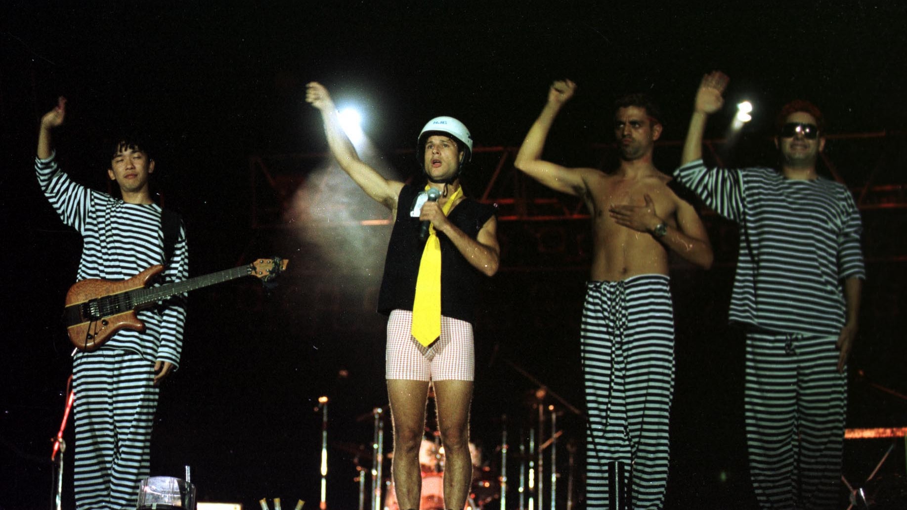 Quatro homens, integrantes da banda Mamonas Assassinas, em palco com figurinos listrados e coloridos, durante show