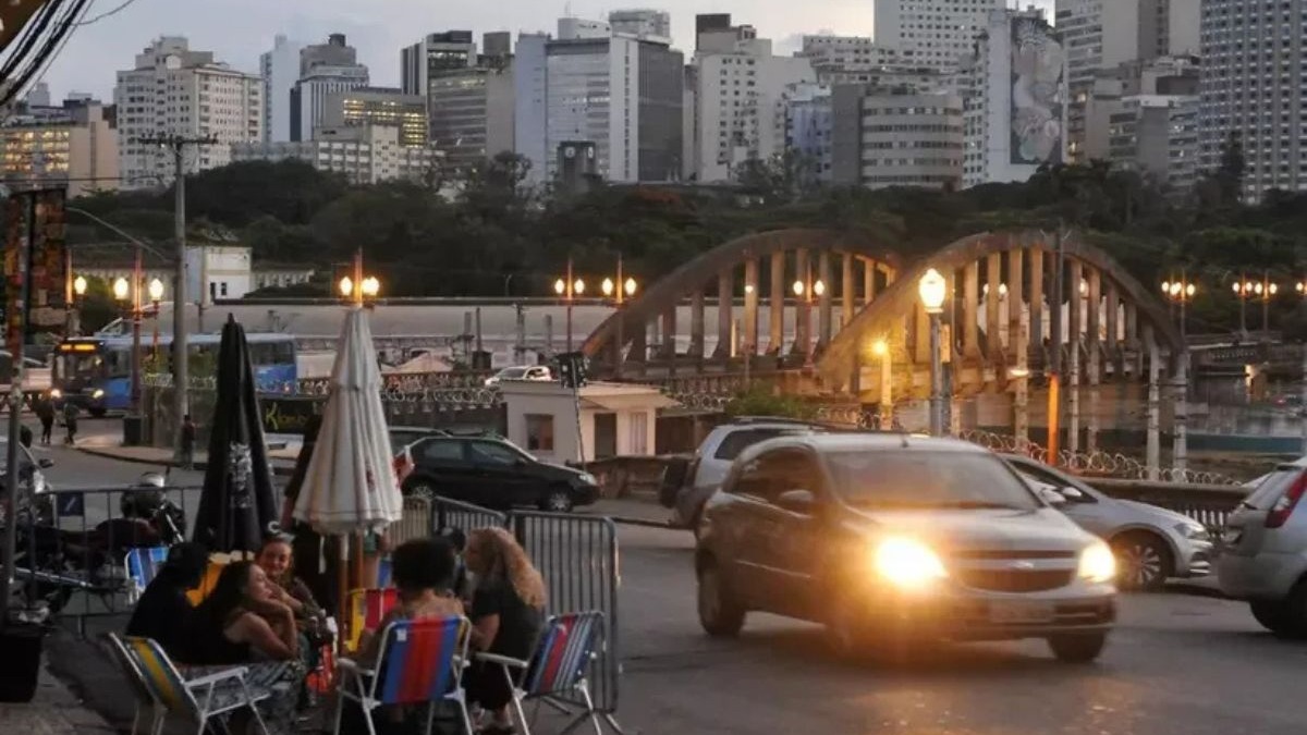 Belo Horizonte, com sua arquitetura e vias planejadas, pulsa entre o presente e o passado que suas ruas e nomes contam. Rua movimentada em Belo Horizonte à noite, pessoas em bar, carros, ponte arqueada e edifícios ao fundo.
