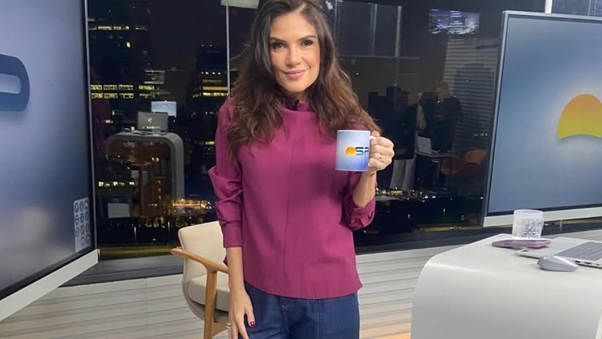 Sabina Simonato em momento de descontração no estúdio do "Bom Dia São Paulo", atração marcada por um clima leve e risadas. Sabina Simonato sorri, segurando caneca com logo SP em estúdio de TV noturno