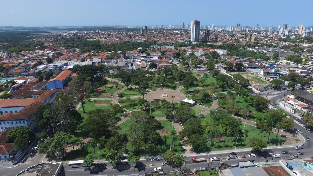 Vista a&eacute;rea de parque urbano verde com ruas, pr&eacute;dios e horizonte de cidade costeira.