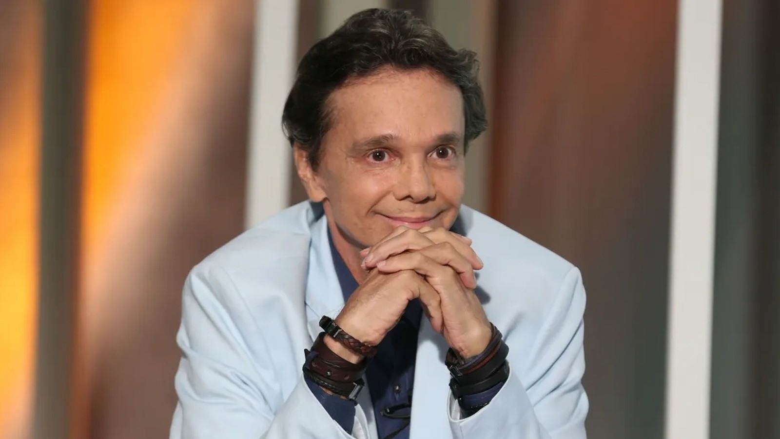 Netinho, ícone do axé music, em imagem que reflete a mensagem de felicidade que deixava aos fãs, antes de seu falecimento aos 60 anos. Cantor Netinho sorri levemente com as mãos cruzadas à frente, vestindo blazer azul claro