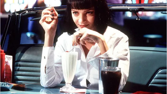 O icônico milkshake de Mia Wallace em Pulp Fiction, inspiração para desvendar os segredos do preparo perfeito da bebida vintage - Foto; Miramax Films Mia Wallace sentada em um estande de lanchonete, segurando milkshake e cigarro, olhando para a câmera