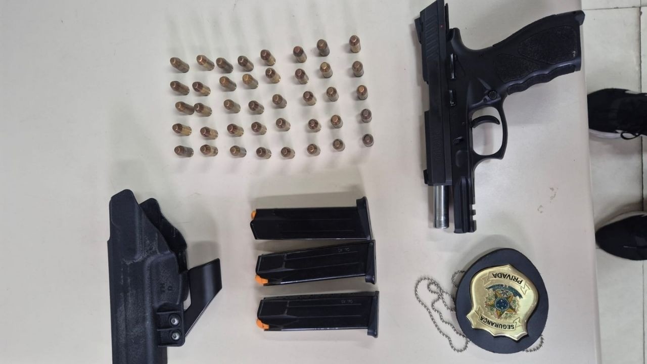 Material apreendido com o homem que se passava por delegado para aplicar golpes em postos de gasolina Pistola preta, três carregadores, dezenas de munições, coldre e distintivo "SEGURANCA PRIVADA" sobre mesa