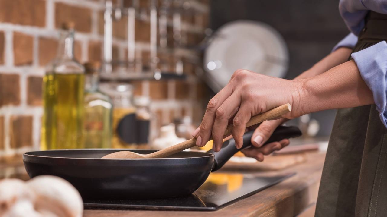 O cozimento da galinhada requer o preparo dos ingredientes em uma panela, etapa essencial para o sabor da receita. Mãos em plano fechado mexem comida em frigideira preta no fogão