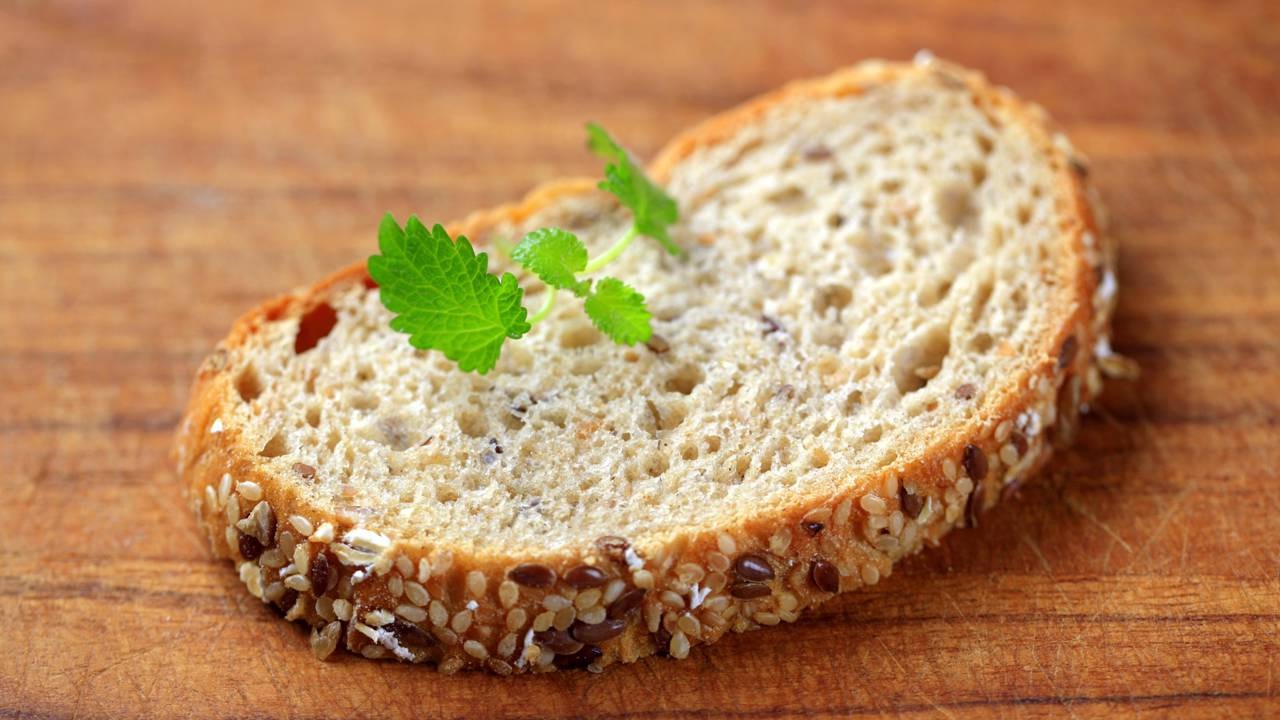 Inicie a semana com um clássico nutritivo: o pão integral é a base de um café da manhã equilibrado para a segunda-feira. Fatia de pão integral com sementes e folha verde sobre tábua de madeira