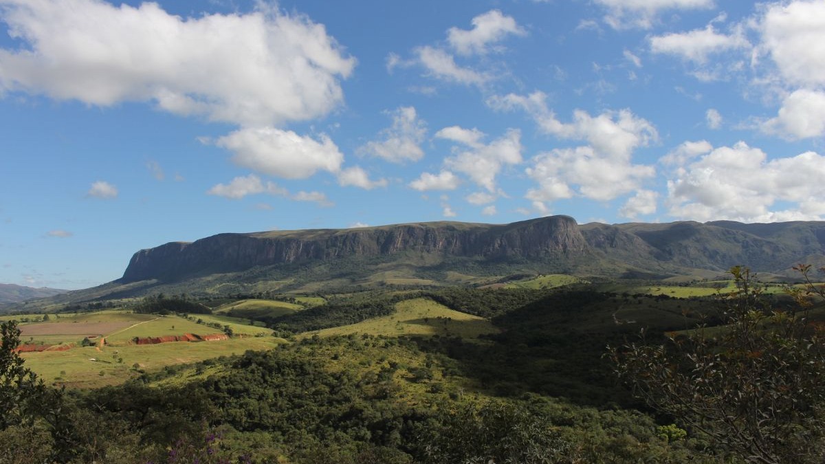 Vista panor&acirc;mica de uma vasta serra com chapada, morros verdes e c&eacute;u azul com nuvens.