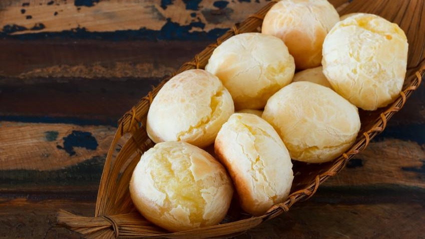 Pães de queijo dourados e com textura ideal, resultado da técnica de escaldar o polvilho abordada na receita. Pães de queijo recém-assados em uma cesta de palha sobre mesa de madeira rústica.