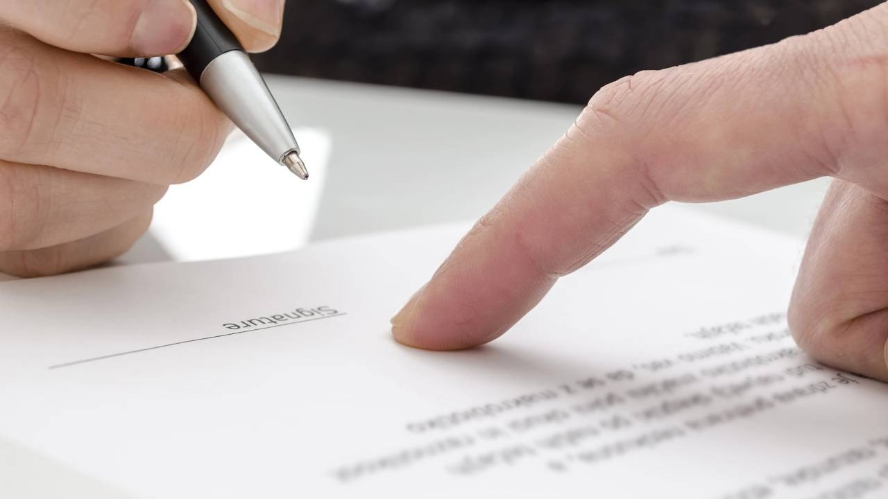 A assinatura de documentos é fundamental para formalizar o testamento e garantir a validade do planejamento sucessório. Mão segurando caneta sobre documento com linha de assinatura, outra mão apontando